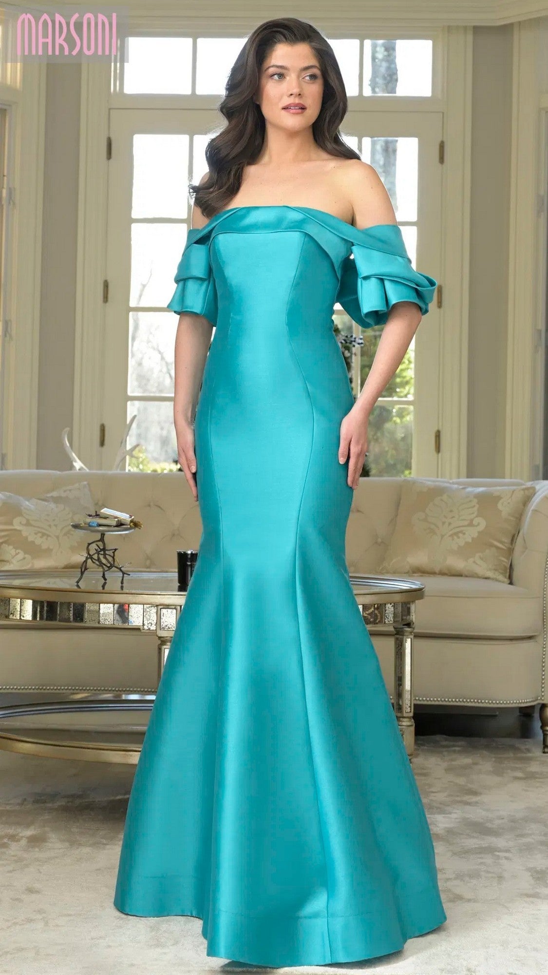 A woman poses indoors in the Marsoni MV1290 long formal dress, featuring an off-the-shoulder fitted mermaid silhouette with ruffled sleeves in Tiffany color, standing before large windows and neutral-toned furniture.